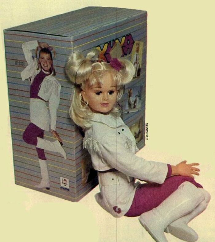 Offensively ugly celebrity doll with blonde hair and white outfit posed sitting next to its original colorful box packaging.