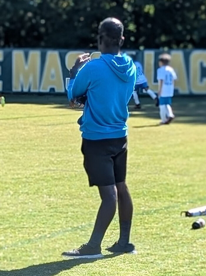Man in blue hoodie and black shorts holding a baseball glove on a sunny field with kids playing in the background unhinged image