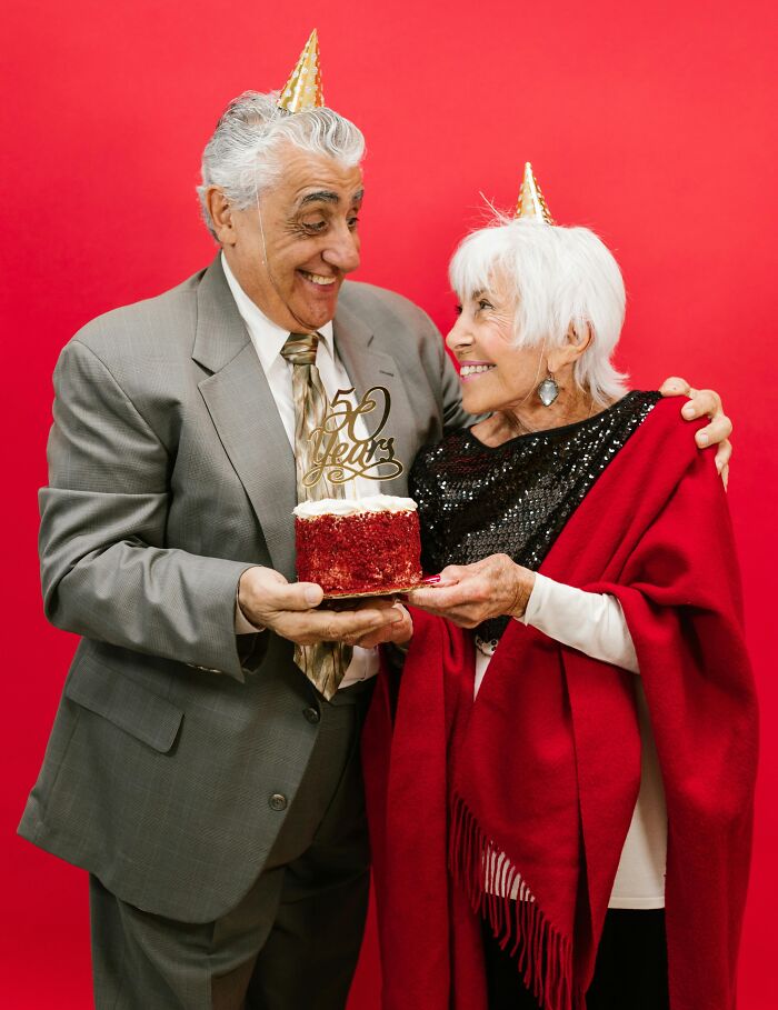 Elderly couple celebrating, smiling at each other with a 50 years cake, wearing party hats against red background.