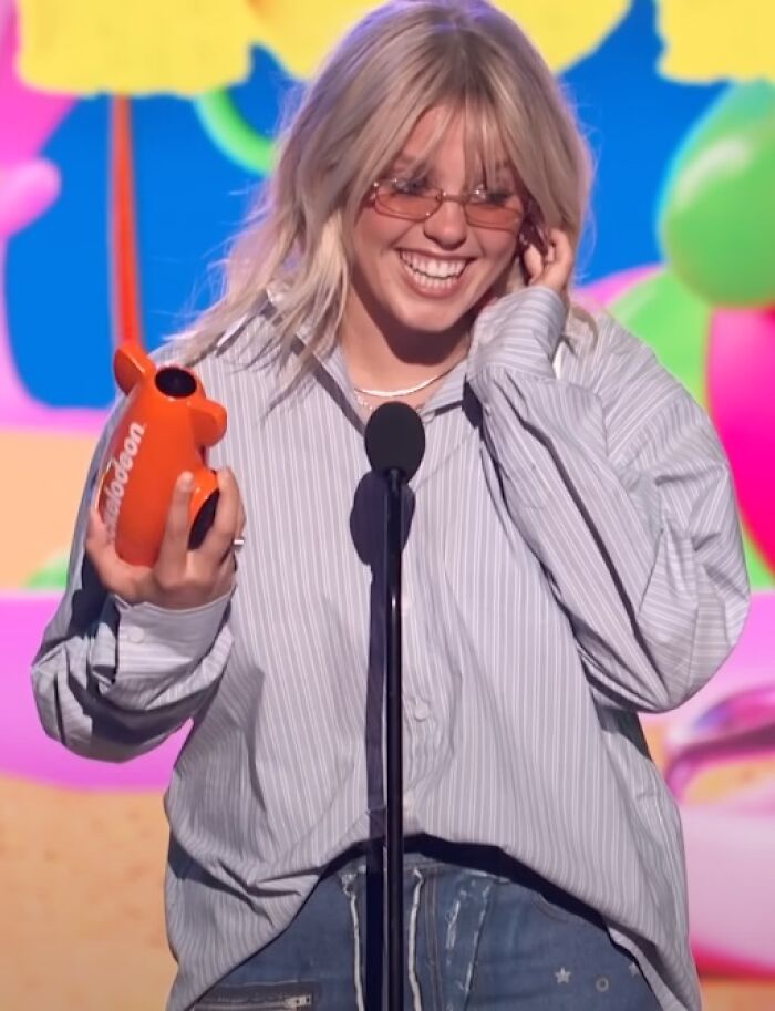 Rene&eacute; Rapp smiling on stage holding an orange award, wearing glasses and a striped shirt at a colorful event.