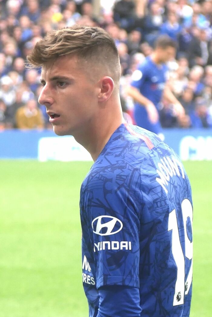 Mason Mount wearing blue Chelsea football kit during a match, focused on the game in a crowded stadium.