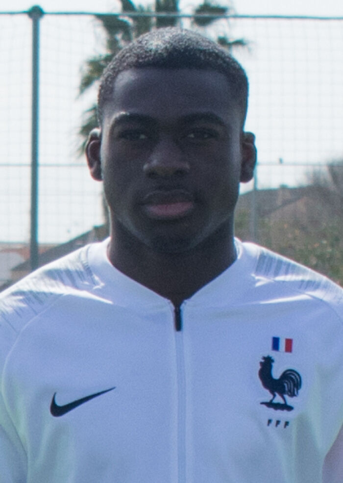 Youssouf Fofana wearing French national team jacket outdoors on a sunny day with sports nets in the background