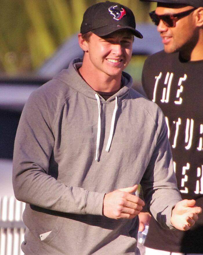 Dylan Edwards smiling outdoors wearing a gray hoodie and black cap, engaging in conversation with another person.