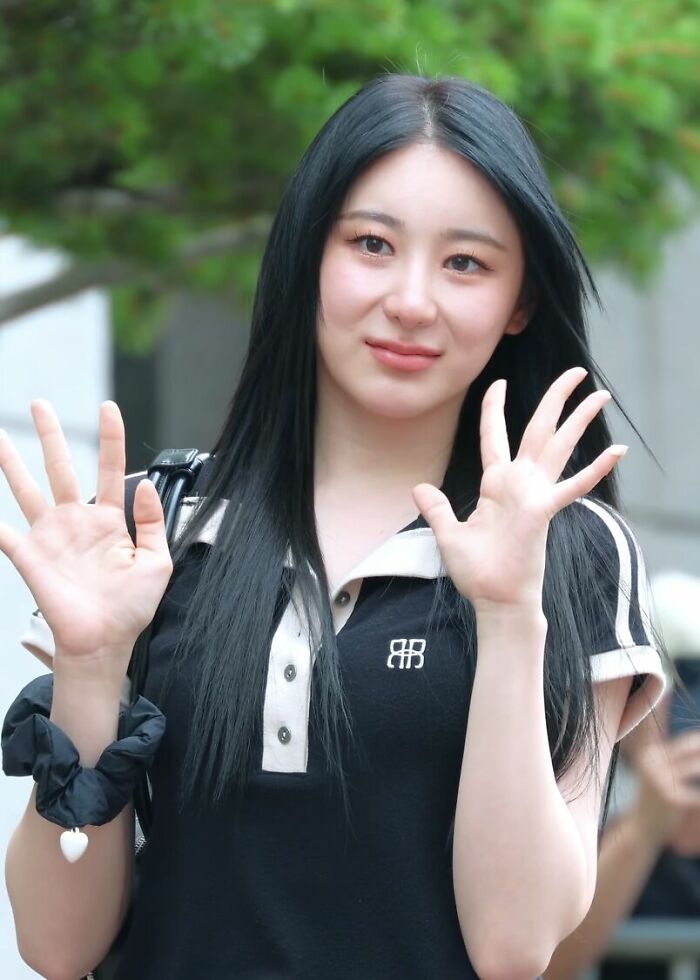 Lee Chae-yeon smiling and waving with long black hair, wearing a black and white shirt outdoors with green background