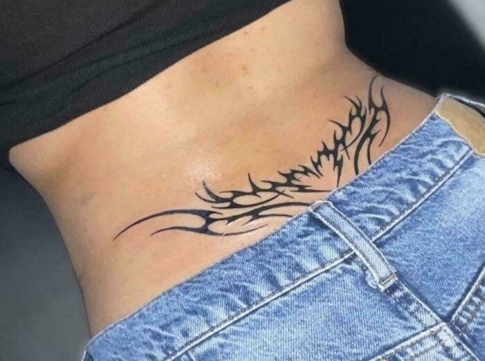 Close-up of old-school tribal tattoo on lower back of person wearing blue jeans and a black top, evoking nostalgia.