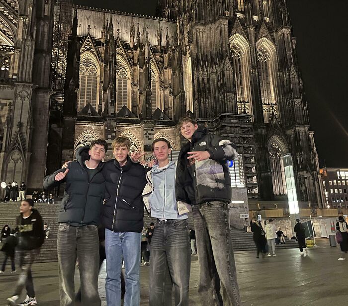 Four tall young men wearing jackets posing together at night in front of a large illuminated Gothic cathedral building.