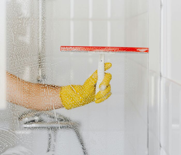 Person wearing yellow gloves cleaning a glass shower door with a red squeegee and soap suds visible.