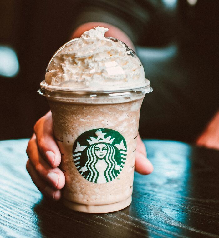 Hand holding a Starbucks Frappuccino with whipped cream on a wooden table, showcasing luxury and rich lifestyle.
