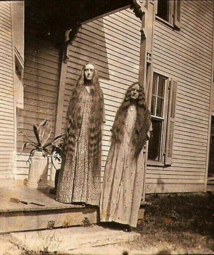 Two women with unusually long hair stand on a porch in a creepy old-school photo that feels slightly cursed.
