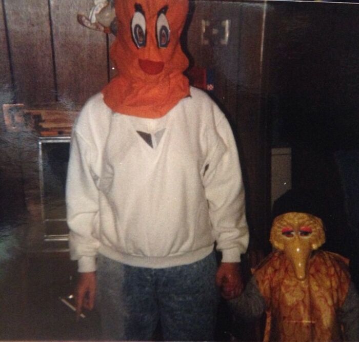 Person in a creepy old-school orange mask holding hands with a child wearing a strange bird costume indoors.