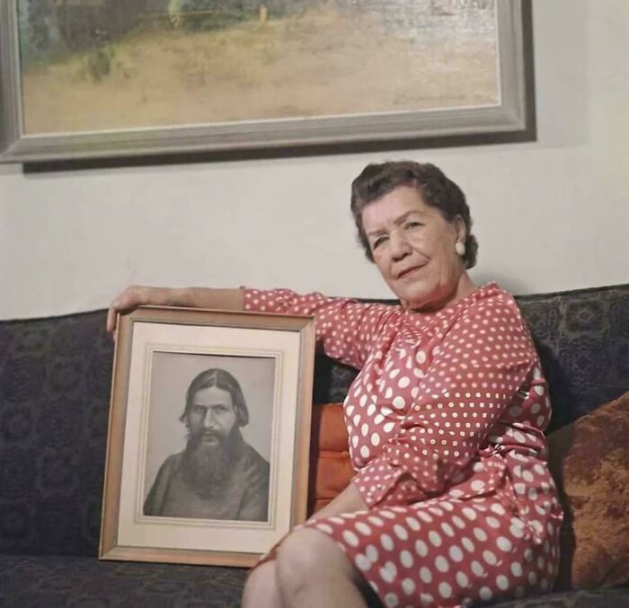 Elderly woman in a polka dot dress sitting with a framed old-school photo that feels slightly cursed on a couch.