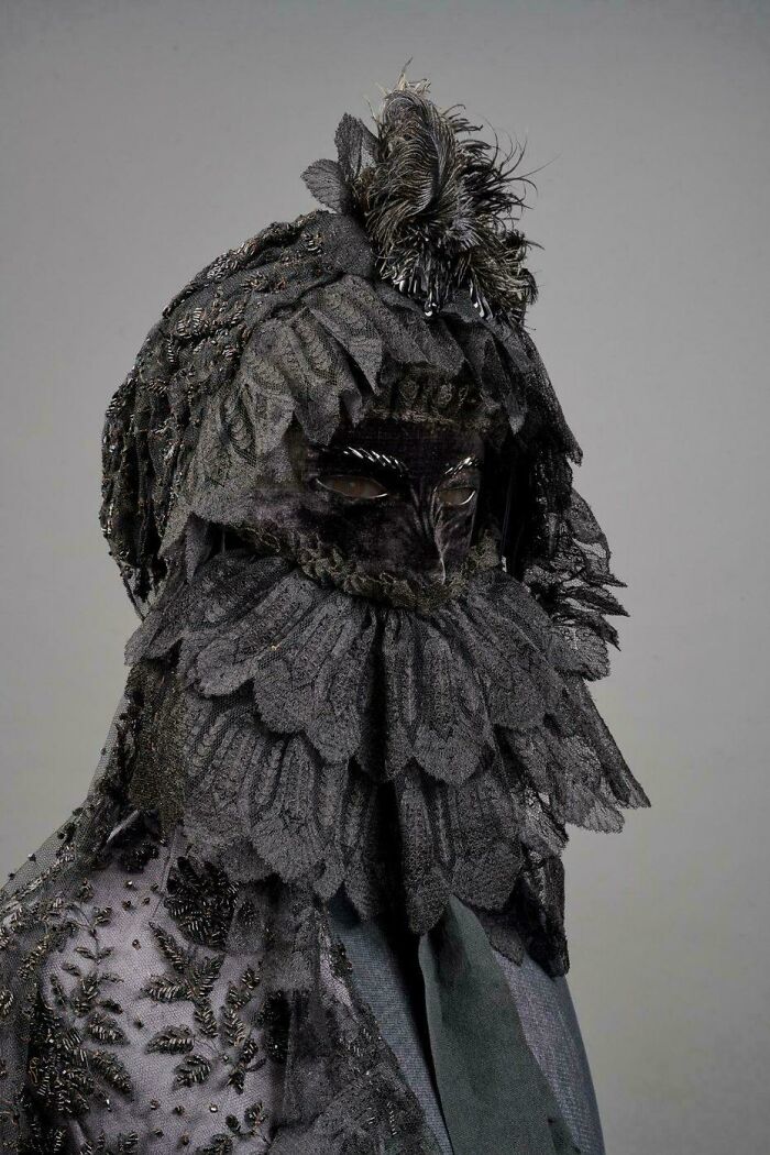 Creepy old-school photo of a person wearing an elaborate black lace mask and costume with feathers and intricate details.