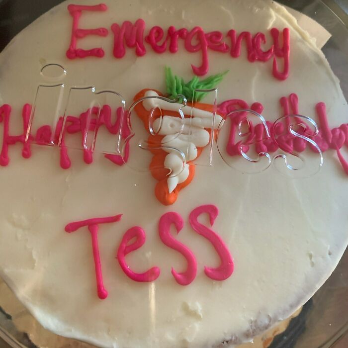 Round white cake with pink icing reading Emergency Happy Birthday Tess and decorated with a small carrot, funny cake photo.