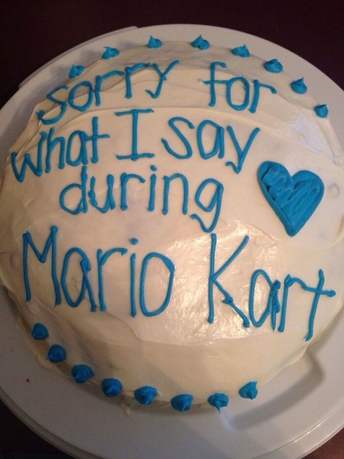 Round white cake with blue icing text apologizing for behavior during Mario Kart, a funny cake showing honest emotions.