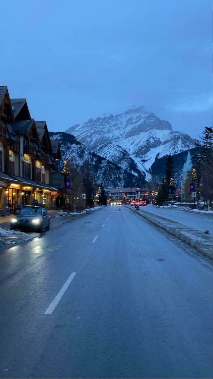 Snowy mountain town street at dusk with cozy lit buildings and cars on a winter evening to enjoy without stepping outside.