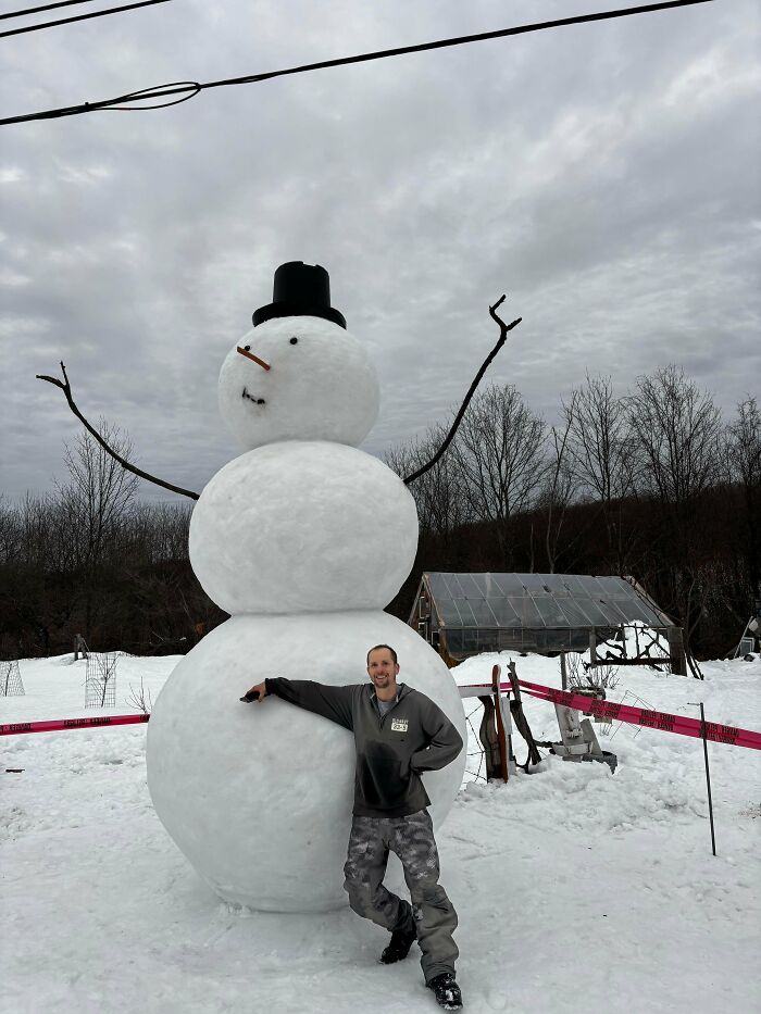 Hombre sonriente junto a un gran muñeco de nieve en un paisaje invernal acogedor con nieve y cielo nublado.