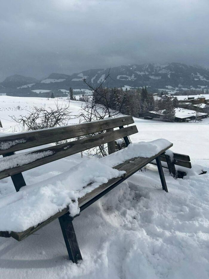 Snow-covered wooden bench in a cozy winter landscape with mountains and trees under a cloudy sky.