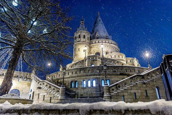Snowfall at night surrounds a cozy lit historic castle with bare trees, perfect for winter enjoyment without stepping outside.