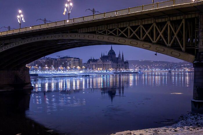 Night view of a cozy winter cityscape with illuminated bridge and calm river reflecting lights under a deep blue sky.