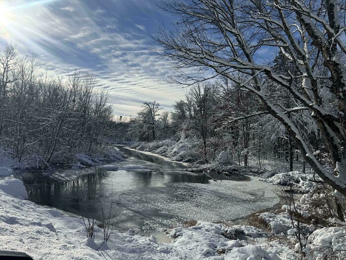 Snow-covered trees and a partially frozen river under a bright sky, showcasing cozy winter nature scenery.
