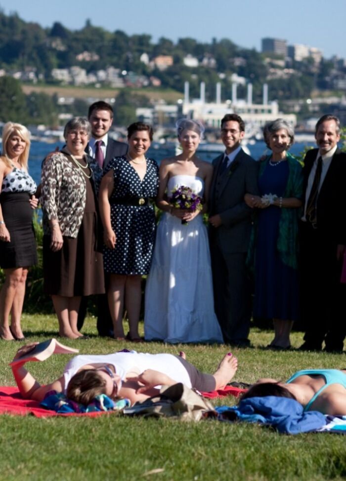 Group posing for a wedding photo by the water while two people sunbathe in the foreground in awkward wedding photos.