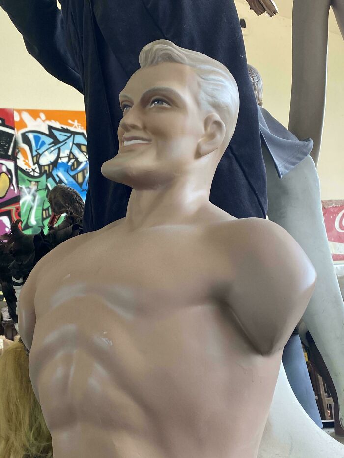 Confusing picture of a smiling headless mannequin torso displayed indoors near graffiti art and other objects.