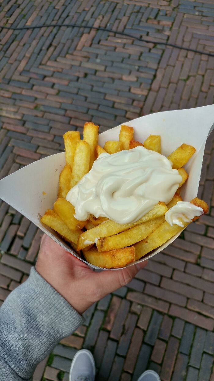 Hand holding a cone of European fries topped with mayonnaise, showcasing normal practices in Europe US may find weird.