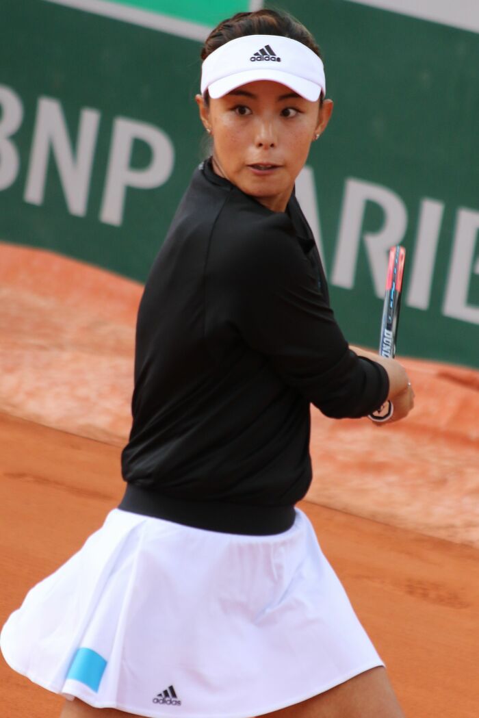Qiang W**g focused on a backhand shot during a clay court tennis match wearing Adidas sportswear.