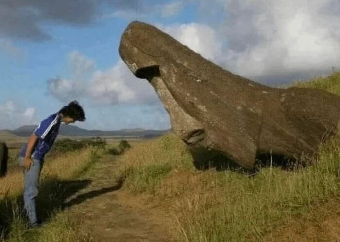 Person bowing to a large Moai statue head on a grassy path, showcasing random and cringe humor moments outdoors.