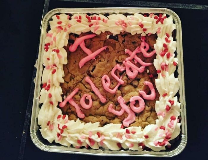 Funny cake with pink icing reading too late joy, decorated with white frosting and red sprinkles in a foil tray.