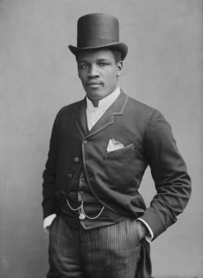 Victorian era man wearing formal suit and top hat, posing confidently with hands in pockets in black and white photo.