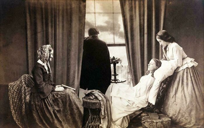 Victorian era family scene with woman seated, man by window, and young woman reclining on a sofa in vintage clothing.