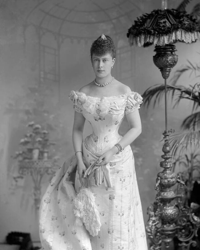 Victorian era woman in elegant floral dress and jewelry standing indoors by ornate lamp and plants.