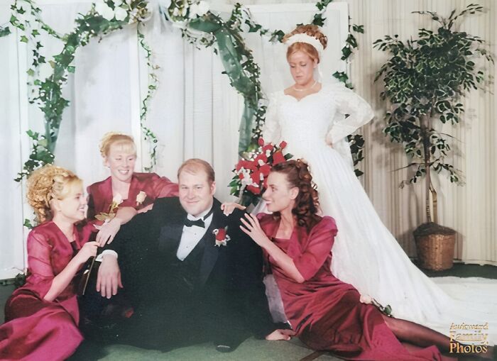 Groom sitting awkwardly on the floor surrounded by bridesmaids in maroon dresses and a bride in a vintage white gown.