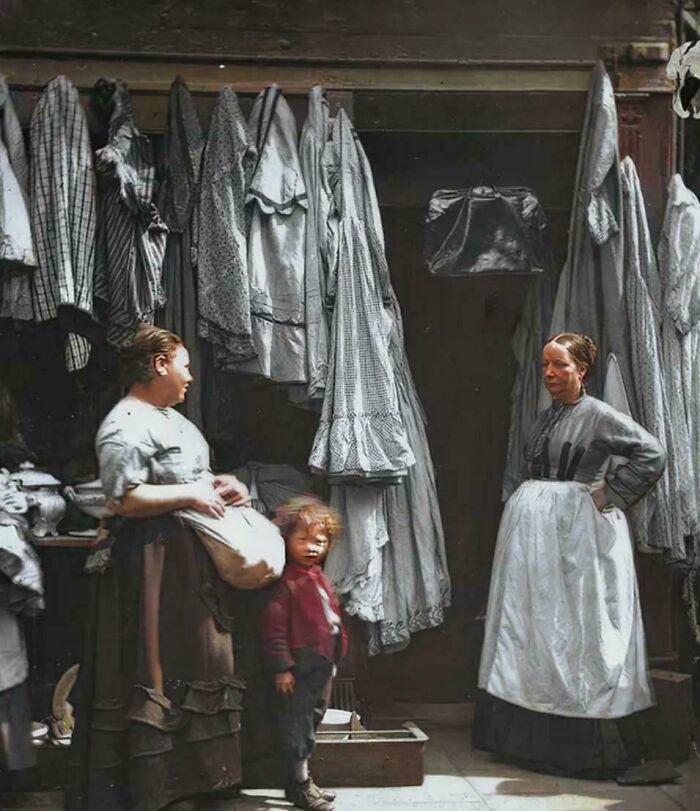 Two women and a child in a Victorian Era laundry setting with hanging clothes, showcasing daily life in the period.