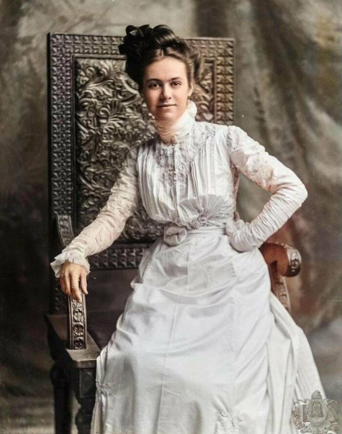 Victorian era woman in a white dress seated on an ornate carved wooden chair, showcasing historic fashion and posture.