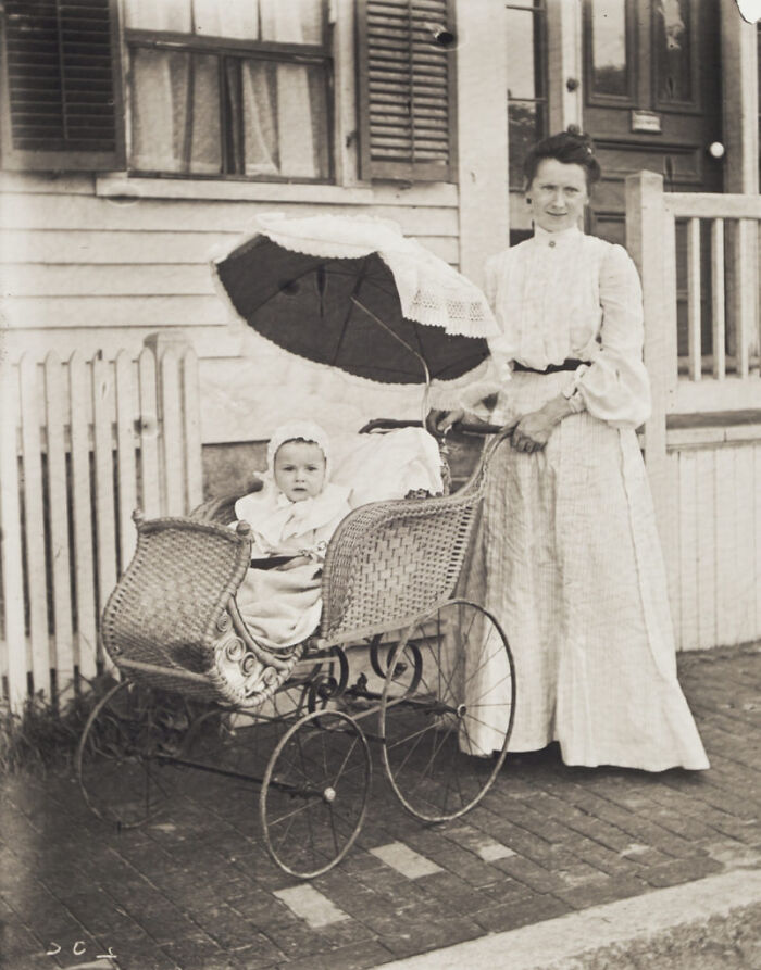 Victorian era mother standing beside woven baby carriage with a child inside on a brick sidewalk outside a house.