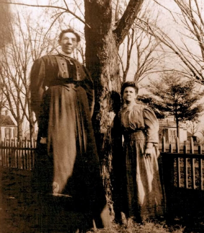 Two Victorian era women standing by a tree, showcasing fashion and lifestyle from the Victorian era in a sepia photo.