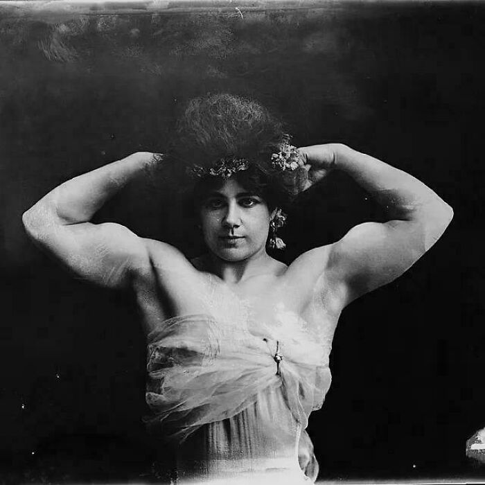 Victorian era strongwoman posing with arms flexed, wearing floral headpiece and sheer fabric garment against dark background.