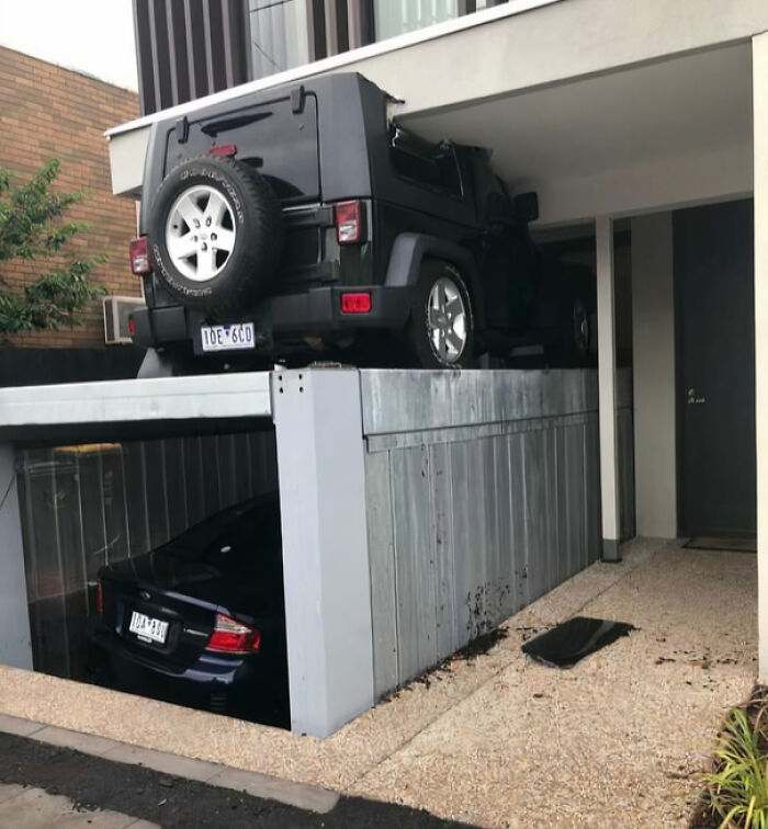 Black SUV stuck on a car lift above another car in a narrow space, showcasing an unfortunate and expensive fail.