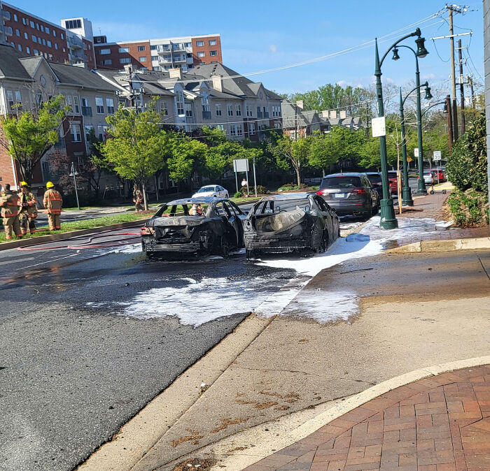 Two burned cars covered in fire extinguisher foam on a city street with firefighters and apartments nearby, costly fail.