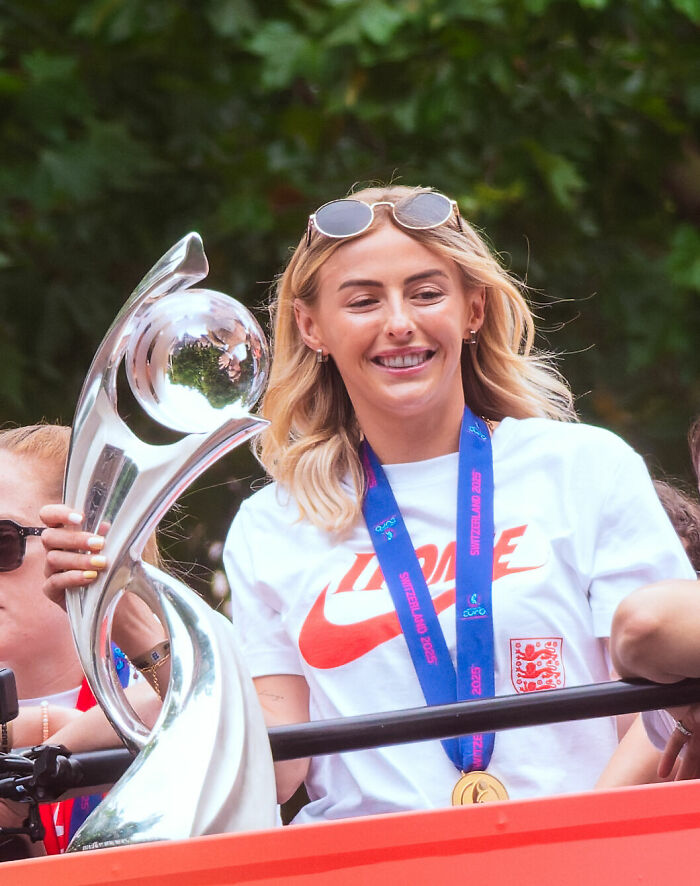 Chloe Kelly smiling and holding a silver trophy, wearing a medal and casual sportswear outdoors.