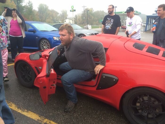 A tall man struggles to get out of a low red sports car while people watch in a parking lot.