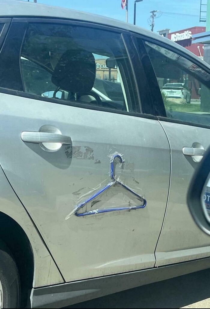 Gray car with a clothes hanger taped to the door as a makeshift handle in a confusing and funny repair attempt.
