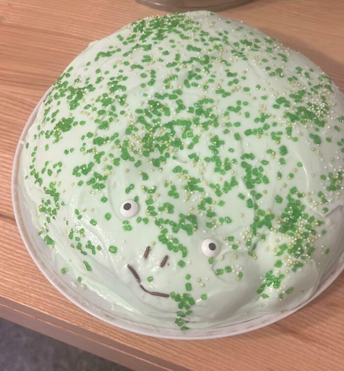 Round cake with uneven pale green frosting and green sprinkles, decorated with candy eyes and a simple face, a dessert fail.