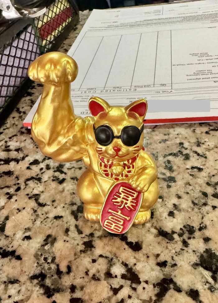 Golden lucky cat figurine with muscular arm and sunglasses sitting on speckled granite counter, a confusing picture.