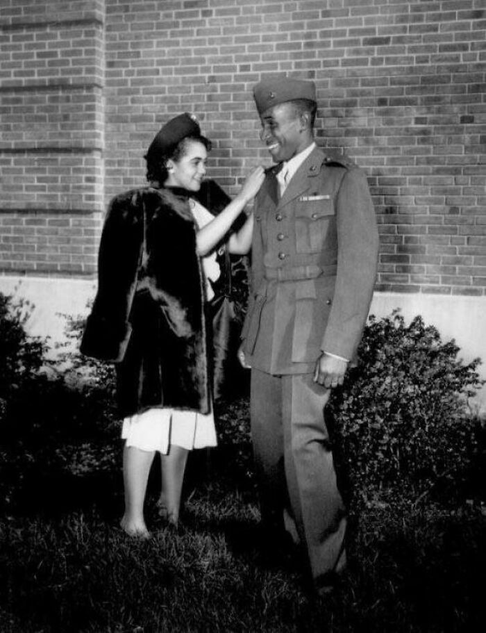 African American soldier in uniform smiling as woman adjusts his collar in historic US history photo.