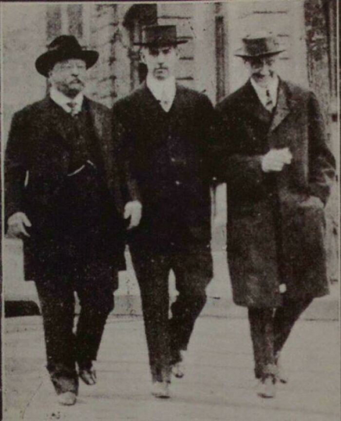 Black and white US history photo showing three men in early 20th century attire walking outside a building.