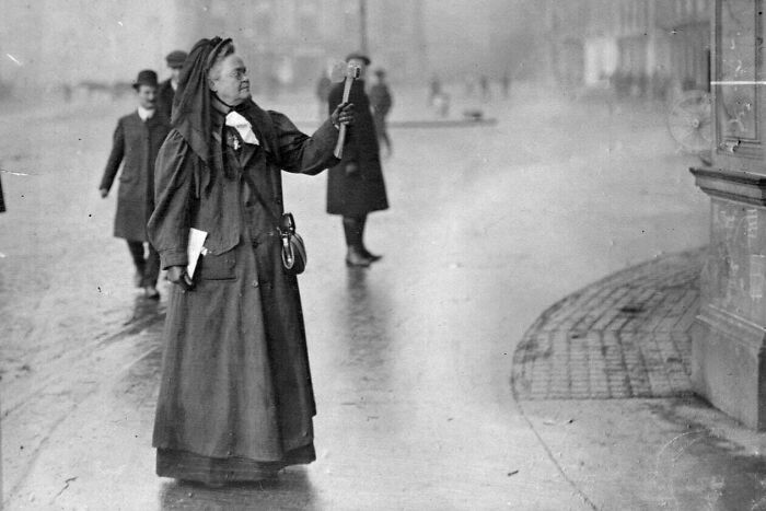 Black and white US history photo showing a woman in period clothing ringing a bell on a city street.