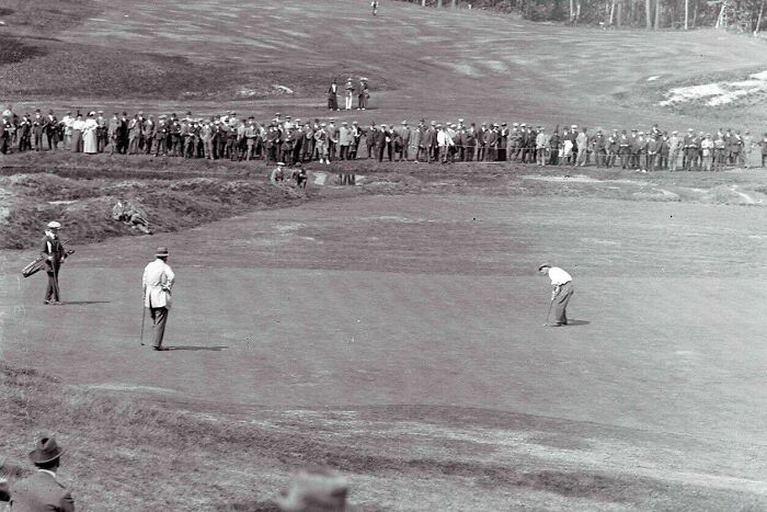 Black and white US history photo showing a group of men playing golf with spectators watching on a grassy field.